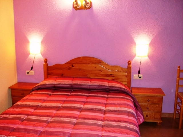 Dormitorio de matrimonio. Pared morada, luz encendida y cabecero de madera